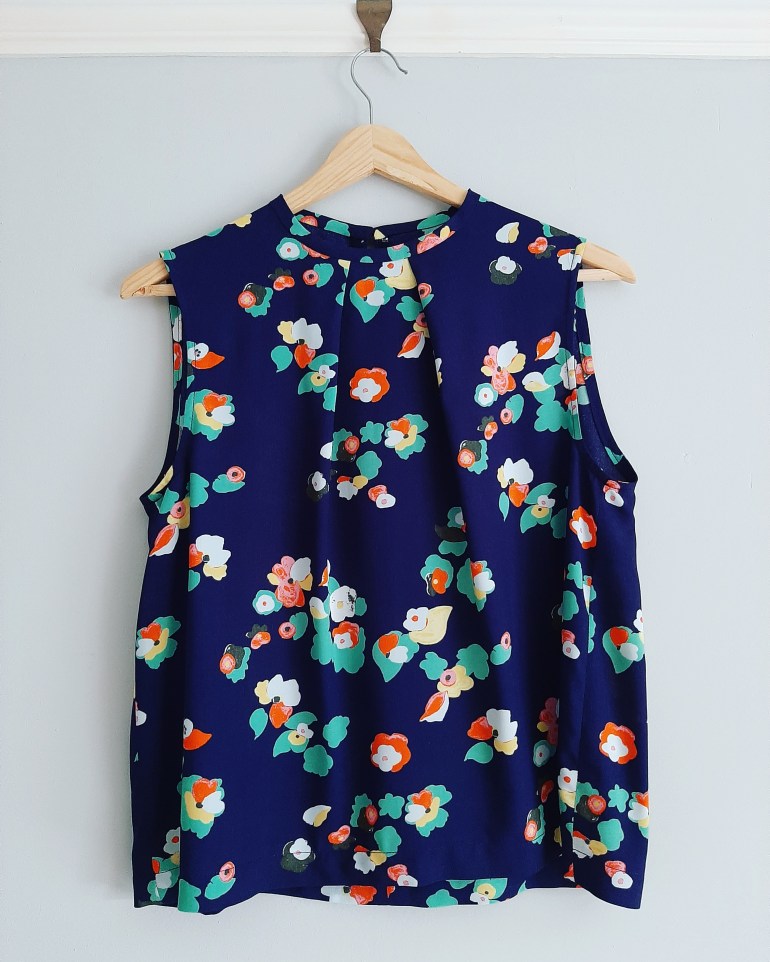 Crew neck sleeveless top with pleated neckline in blue and multicoloured floral viscose fabric hangs on wall