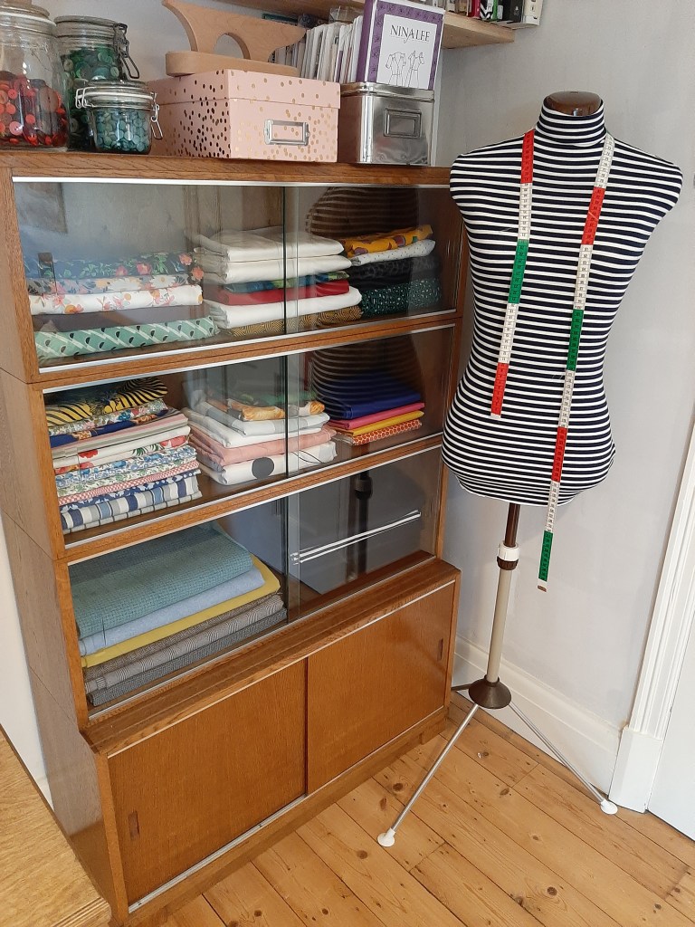 A glass-fronted sewing cabinet repurposed to hold neatly folded fabric stash is in the corner of a room next to a dressmaker's mannequin in a stripe cover with a tape measure hanging from its neck. Above, patterns are stored with a tailor's clapper and kilner jars filled with buttons.